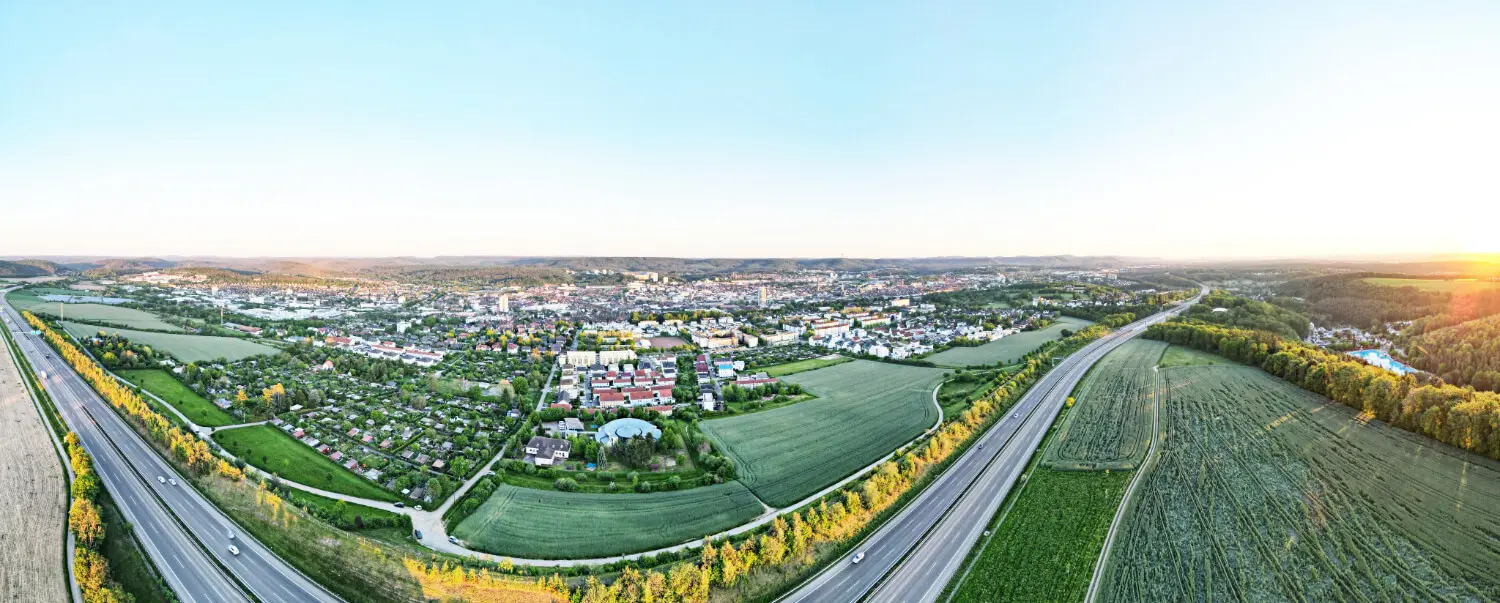 Banner der Webseite. Drohnenaufnahme. Zu sehen ist die Autobahn A6, im Hintergrund die Stadt Kaiserslautern, die Sonne ist am untergehen. Insgesamt sehr viel grün.